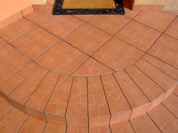 Wyłożone terakotą schody wejściowe z zaokrąglonym stopniem i wycieraczką przed drzwiami, widoczne z góry, w słoneczny dzień.