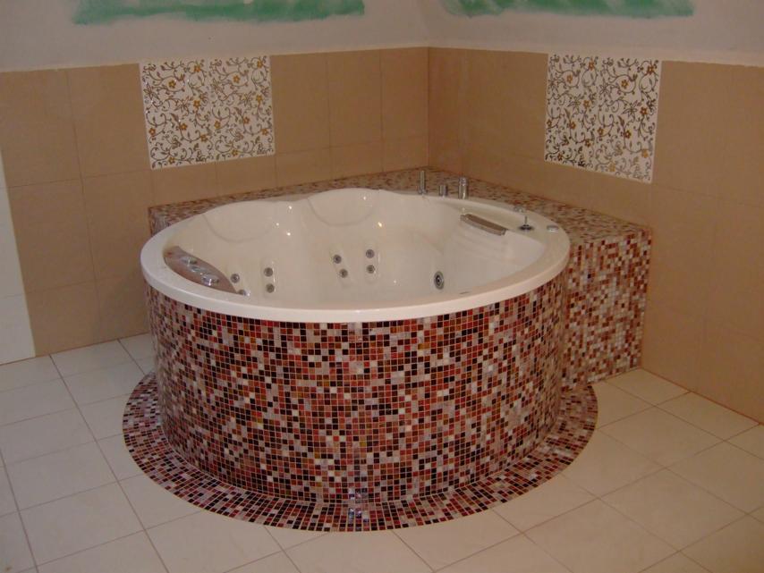 Okrągła wanna z hydromasażem obłożona mozaiką w odcieniach brązu i czerwieni, z ozdobnymi płytkami na ścianie w tle.