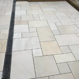 Ułożona jasna kostka brukowa z odpływem liniowym, widoczne plastikowe krzyżyki dystansowe, buty robocze w prawym górnym rogu.