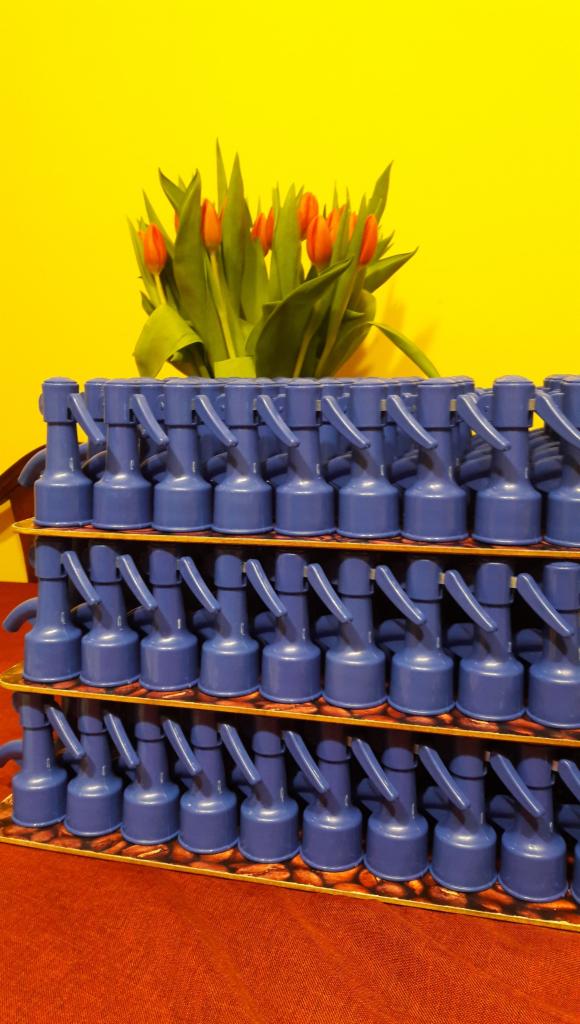 Rzędy niebieskich, plastikowych zaworów na tekturowej tacy z motywem kawy, w tle bukiet czerwonych tulipanów na żółtym tle.