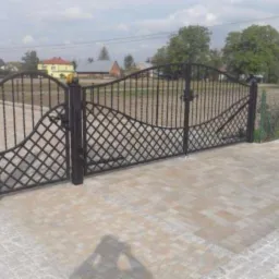 Czarna, metalowa brama wjazdowa z dekoracyjnym wypełnieniem w romby, zamykana na zamek, na tle domów i drzew.