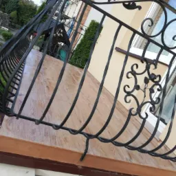 Balkon z kutą, czarną balustradą o zdobieniach roślinnych, wykończony płytkami imitującymi drewno, widok z dołu na tle elewacji budynku.