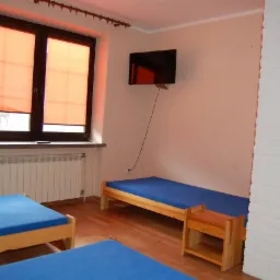 Pokój z dwoma łóżkami z niebieskimi materacami, drewnianą podłogą, telewizorem na ścianie i oknem z pomarańczowymi roletami, ściana wykończona białą cegłą.