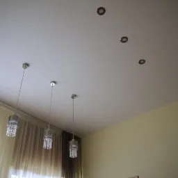 Biały sufit z trzema lampami wiszącymi z kryształkami i czterema punktowymi oprawami oświetleniowymi wpuszczonymi, widok z dołu, zasłony w tle.