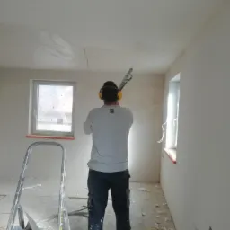 Mężczyzna w słuchawkach ochronnych aplikuje tynk maszynowy na sufit w pomieszczeniu w trakcie prac wykończeniowych, widoczna drabina malarska i resztki materiałów na podłodze.
