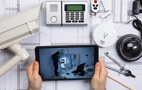 Plan instalacji zabezpieczeń z elementami systemu: kamera zewnętrzna, centrala alarmowa, czujnik dymu, kamera kopułkowa; na tablecie widoczny podgląd z kamery z włamywaczem w biurze.