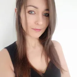 Portret kobiety z długimi brązowymi włosami i niebieskimi oczami, ubrana w czarną sukienkę na tle białej ściany.