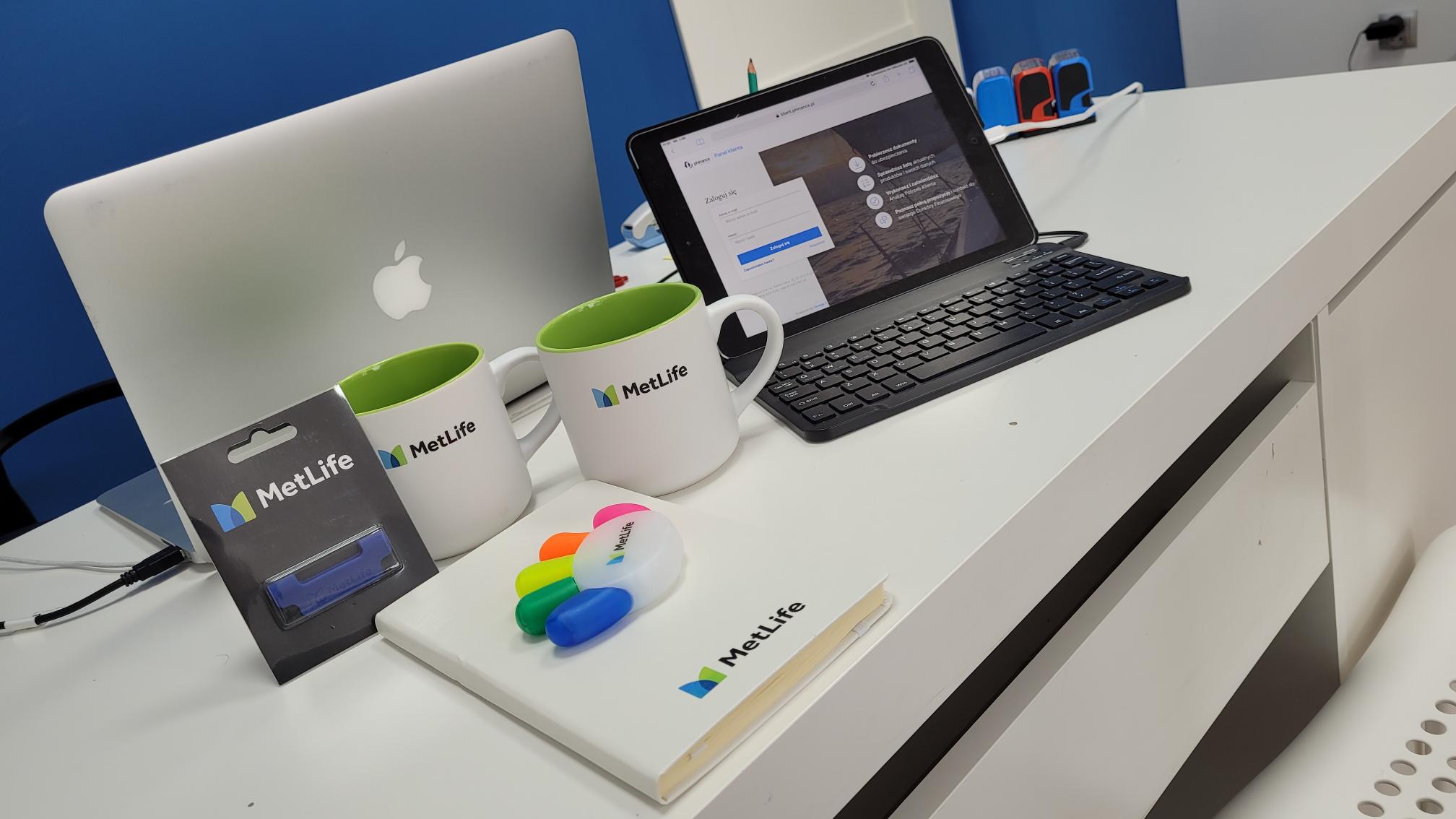Biurko z laptopem Apple, tabletem z otwartą stroną logowania, dwoma kubkami z logo MetLife, notatnikiem, zakreślaczem w kształcie dłoni i opakowaniem z długopisem. Niebieska ściana w tle.