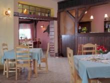 Rustykalne wnętrze restauracji z drewnianymi elementami konstrukcyjnymi, stołami nakrytymi błękitnymi obrusami i bukietami czerwonych kwiatów na stolikach.