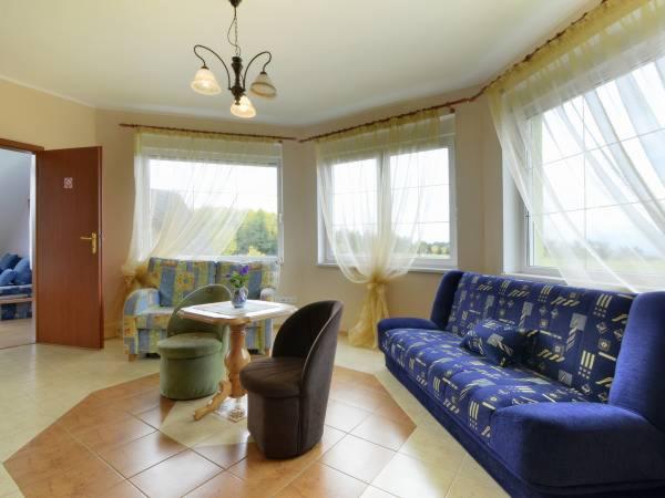 Jasne wnętrze pokoju dziennego z niebieską sofą, stolikiem i krzesłami, widok na zieleń za oknem.