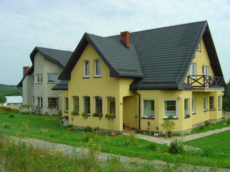 Dwa domy jednorodzinne: żółty z balkonem i numerem 69 oraz szary, otoczone zielonym trawnikiem z kwiatami w doniczkach.