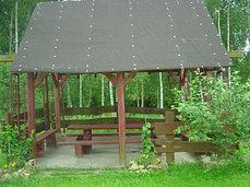 Drewniana altana z ciemnym dachem w ogrodzie, widoczne ławki i stół, tło stanowią drzewa liściaste.