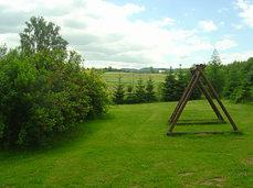 Drewniana konstrukcja huśtawki na trawniku, w tle łąka i drzewa, pochmurne niebo.