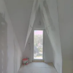 Poddasze ze skośnymi ścianami i oknem w szczycie, ściany przygotowane do malowania, widoczne ślady szpachlowania, na podłodze stoi wiadro z masą szpachlową.