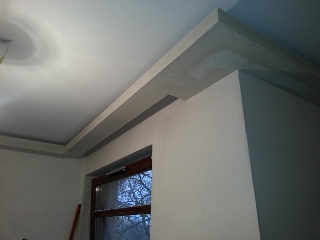 Wykończony sufit z płyt kartonowo-gipsowych z widocznym oknem i fragmentem drabiny w rogu pomieszczenia.