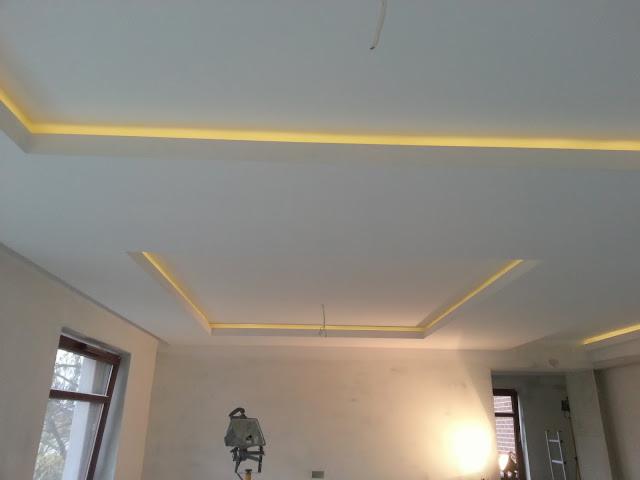 Sufit z podwieszanymi elementami i oświetleniem LED w trakcie prac wykończeniowych, widoczne okno i drabina w tle.