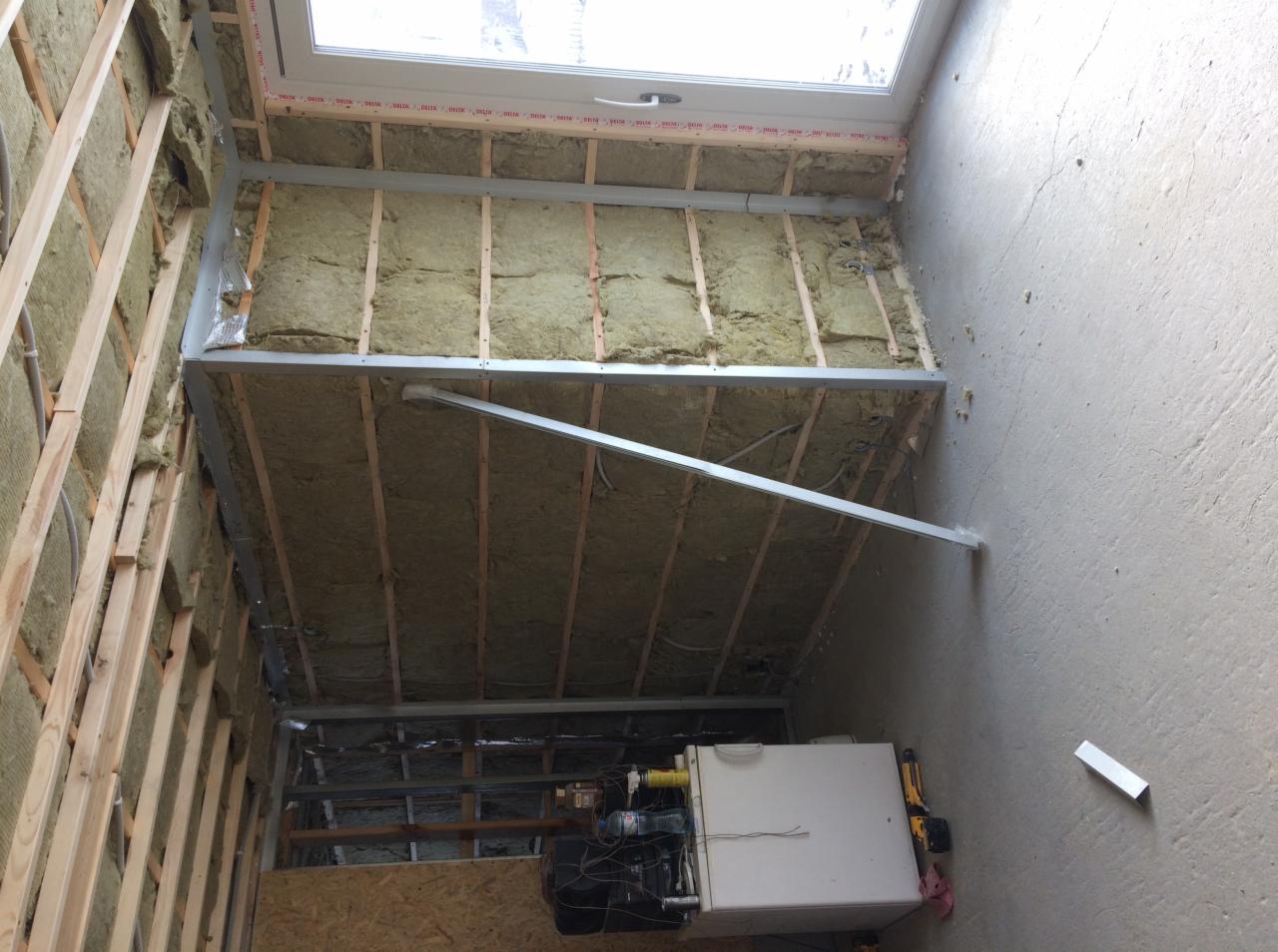 Wnętrze w trakcie prac: widoczna izolacja z wełny mineralnej między drewnianymi belkami na ścianie i suficie, metalowe profile, okno dachowe, instalacja elektryczna i fragment kotła grzewczego.