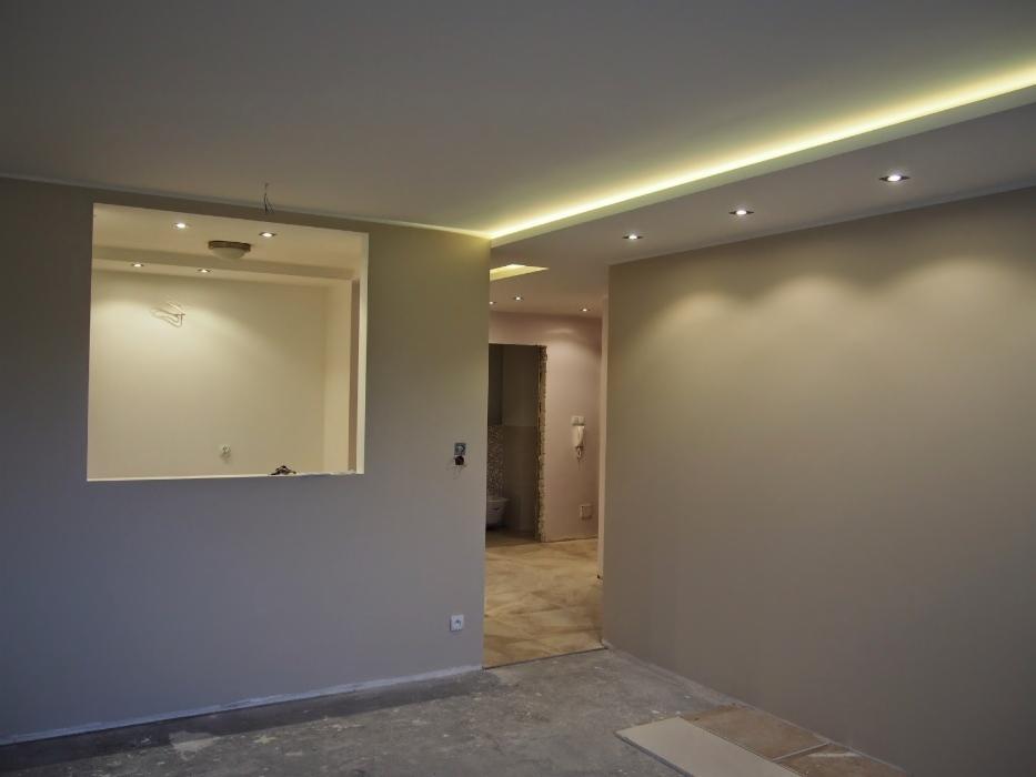 Wnętrze w trakcie prac wykończeniowych: widok na ścianę z wnęką oświetloną punktowo, sufit z listwą LED, podłoga w stanie surowym.