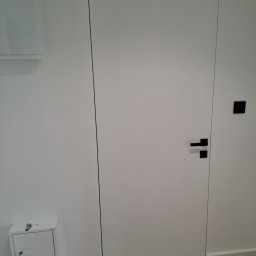 TarBud - Białe, minimalistyczne drzwi z czarną klamką i ukrytą ościeżnicą, zamontowane w świeżo odmalowanej, białej ścianie, z widoczną skrzynką z kluczem poniżej i brązową podłogą z płytek imitujących drewno.
