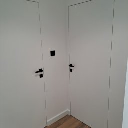 TarBud - Dwa białe, minimalistyczne drzwi wewnętrzne z czarnymi klamkami, ukryte w ścianie, widoczne na tle drewnopodobnej podłogi i białych listew przypodłogowych.