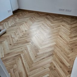 TarBud - Ułożona parkietowa podłoga w jodełkę, widoczne gniazdka elektryczne na ścianie, fragment grzejnika i rolki materiału w rogu pomieszczenia.