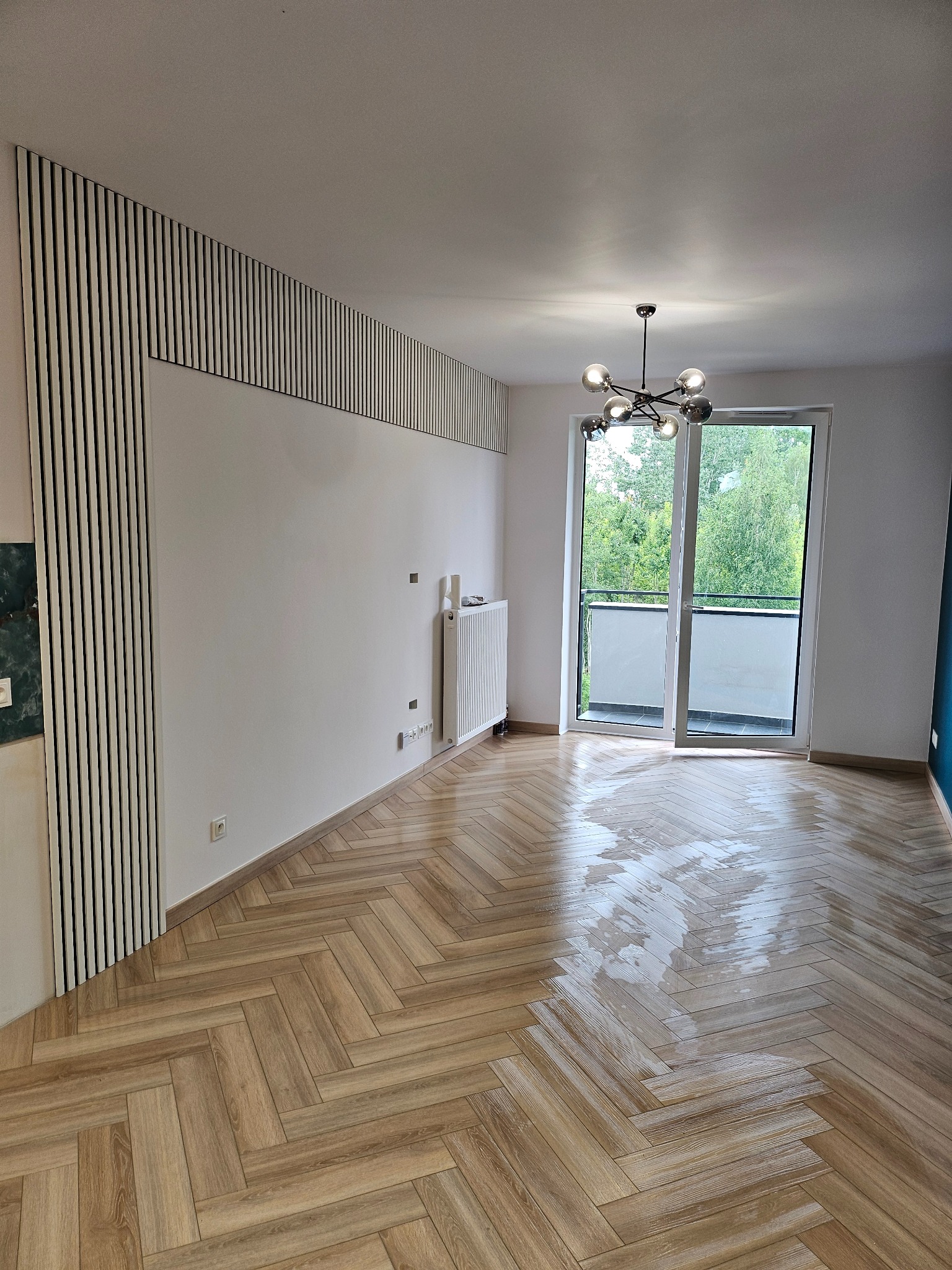 Jasne, przestronne wnętrze po remoncie z parkietem ułożonym w jodełkę, białymi ścianami z dekoracyjnymi pionowymi listwami, nowoczesną lampą sufitową i wyjściem na balkon.