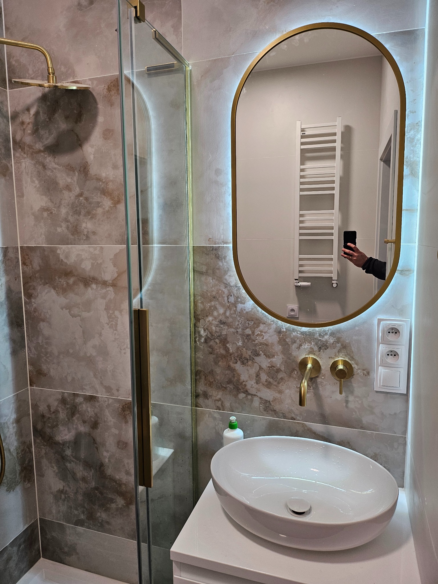 Nowoczesna łazienka z kabiną prysznicową ze złotymi elementami, umywalką nablatową i podświetlanym lustrem owalnym, odbijającym biały grzejnik drabinkowy.