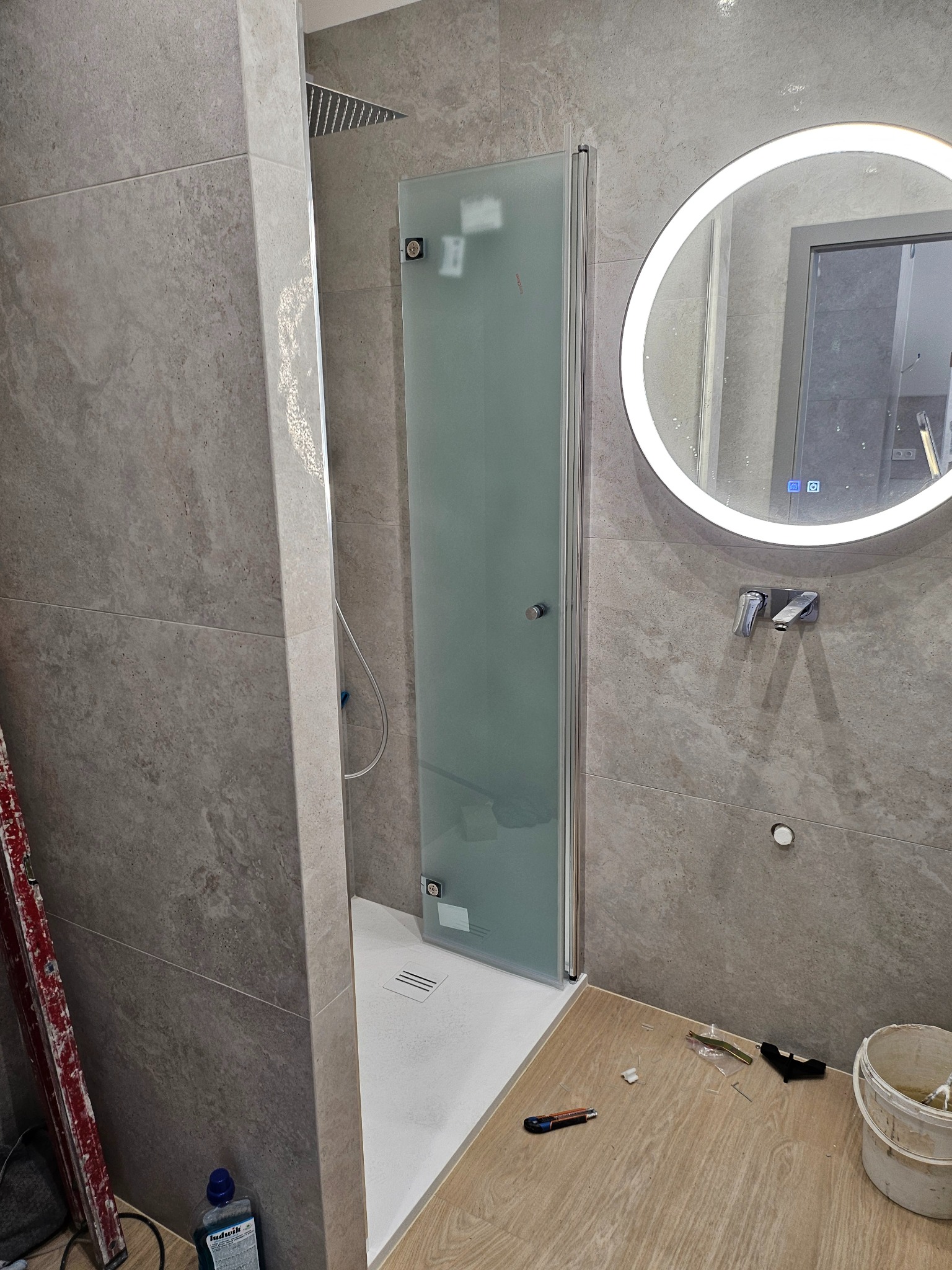 Wykończona łazienka z kabiną prysznicową z matowego szkła, okrągłym lustrem podświetlanym LED, szarymi płytkami imitującymi kamień na ścianach i drewnopodobną podłogą. Widoczne akcesoria montażowe...