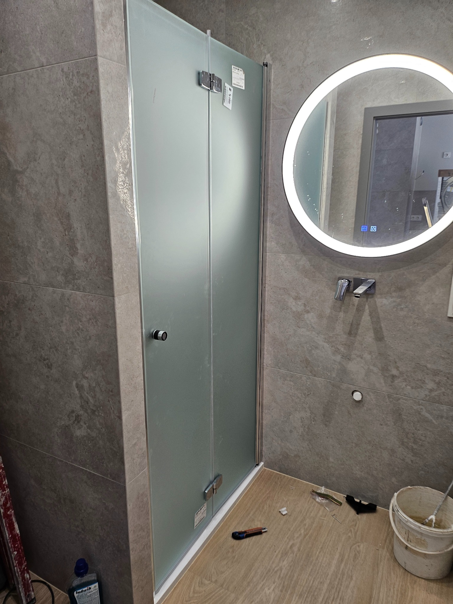 Nowa, matowa kabina prysznicowa zamontowana w łazience wyłożonej płytkami imitującymi kamień, okrągłe lustro z podświetleniem LED, widoczne narzędzia i wiadro po fugowaniu.