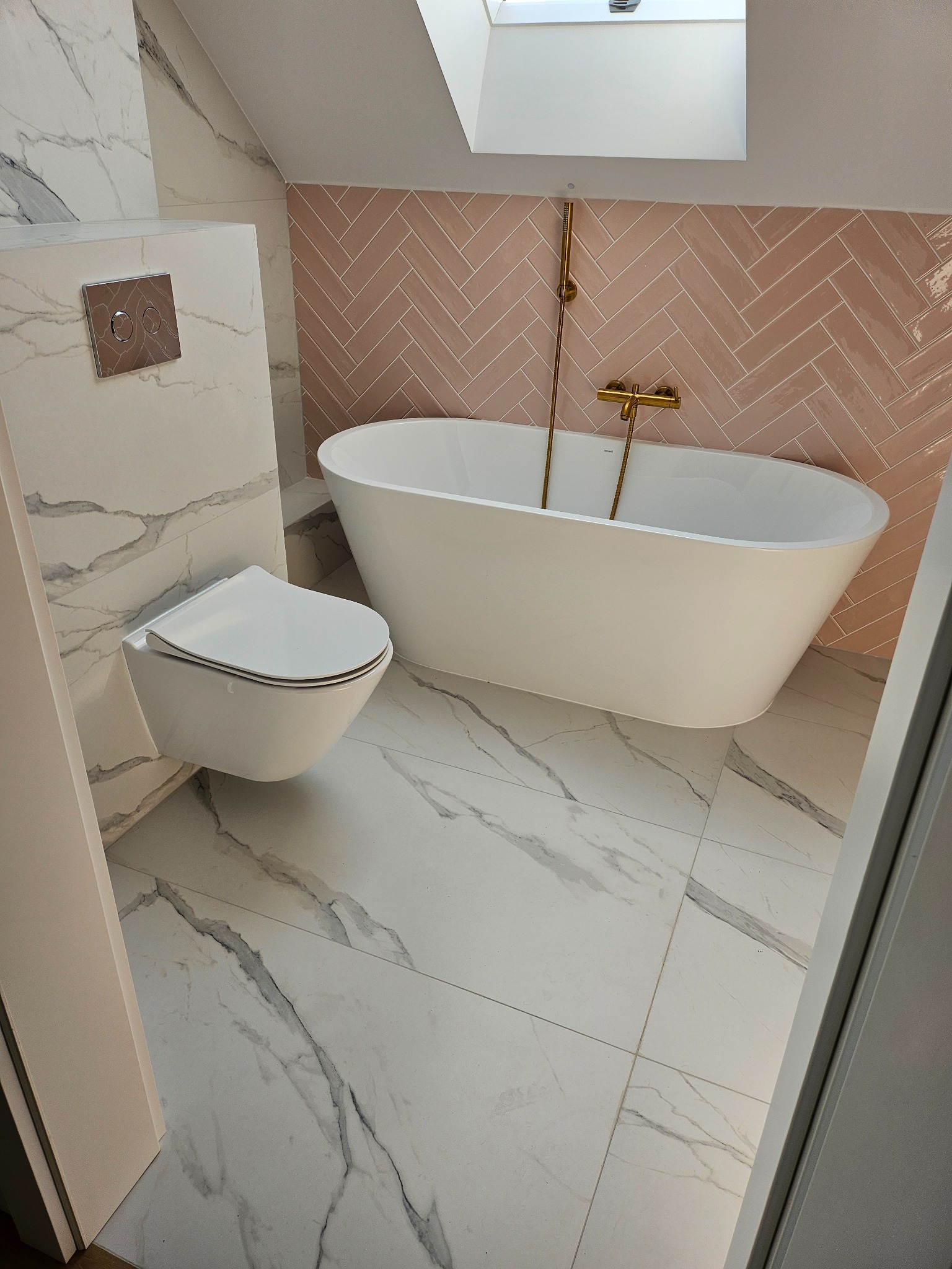 Nowoczesna łazienka na poddaszu z białą armaturą, marmurowymi płytkami podłogowymi i ściennymi oraz różową ścianą z płytek ułożonych w jodełkę, oświetlona światłem wpadającym przez okno dachowe.