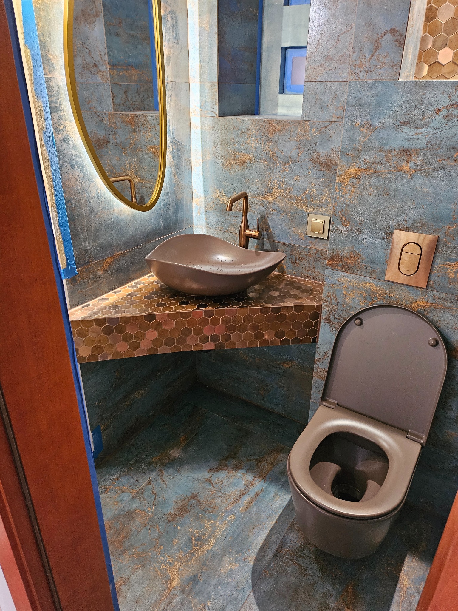 Nowoczesna łazienka z umywalką w kształcie misy, armaturą w kolorze brązu i płytkami w odcieniach niebieskiego i brązu, z widocznym fragmentem toalety i lustra z podświetleniem LED.