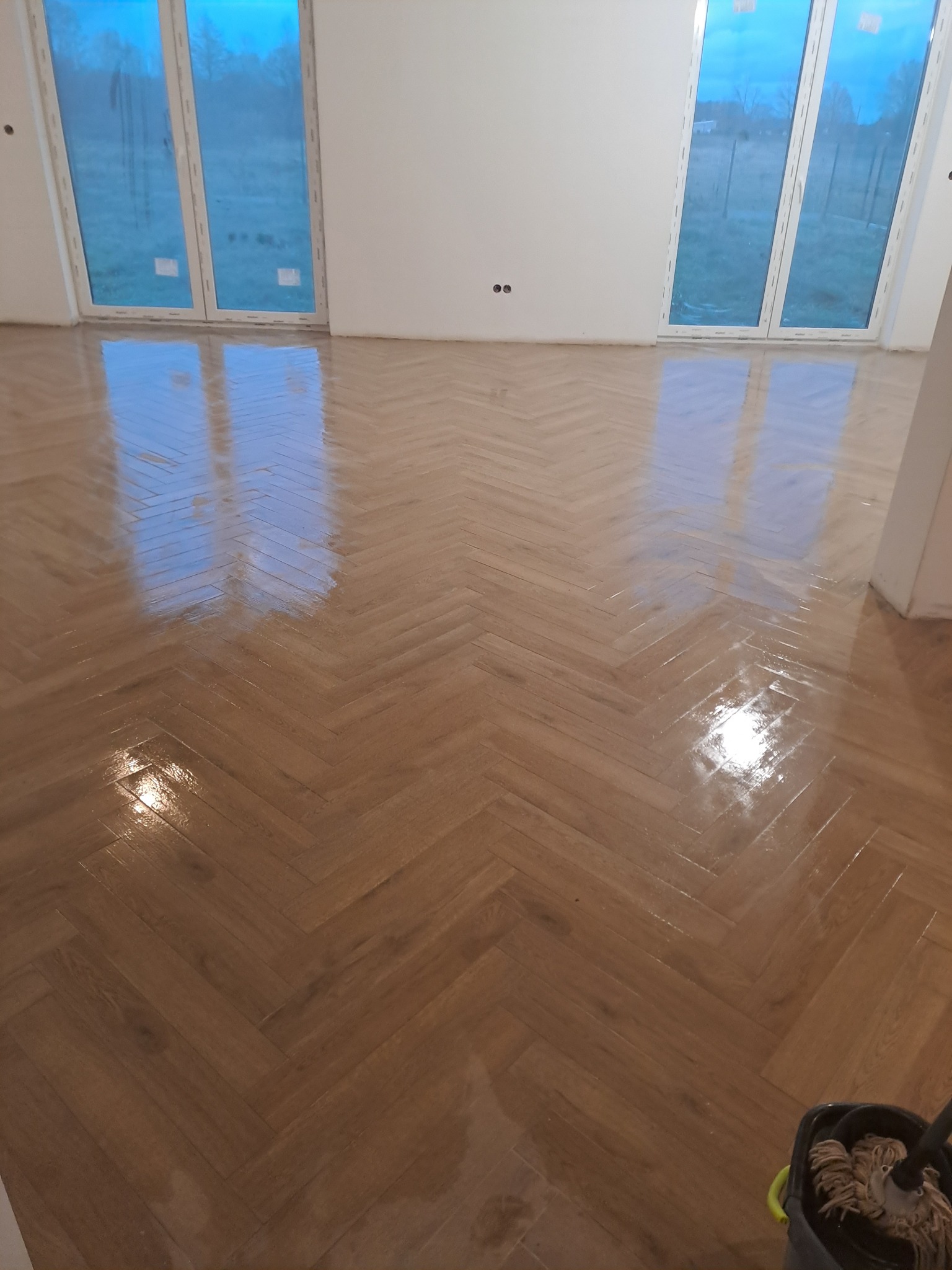 Świeżo ułożona, błyszcząca podłoga z paneli ułożonych w jodełkę, z widocznym odbiciem okien i wiadrem z mopem w rogu.