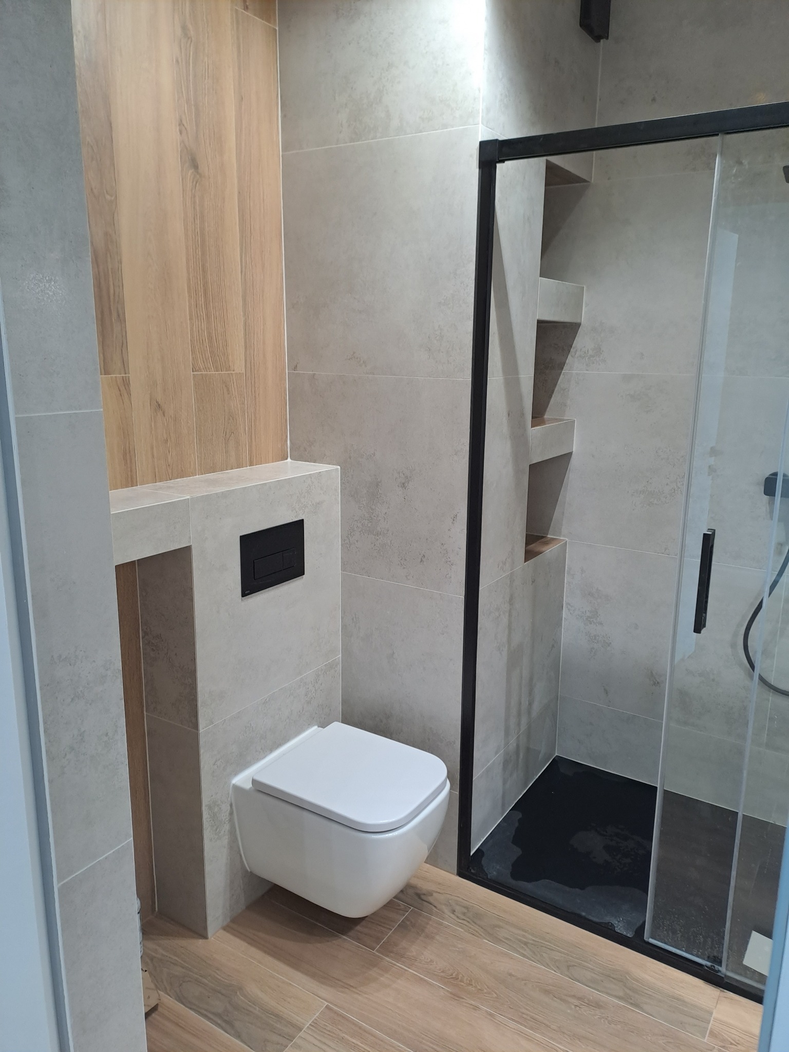 Nowoczesna łazienka z płytkami imitującymi drewno na podłodze i częściowo na ścianie, szare płytki na pozostałych ścianach, wisząca toaleta z czarnym przyciskiem spłukującym oraz kabina prysznicowa...