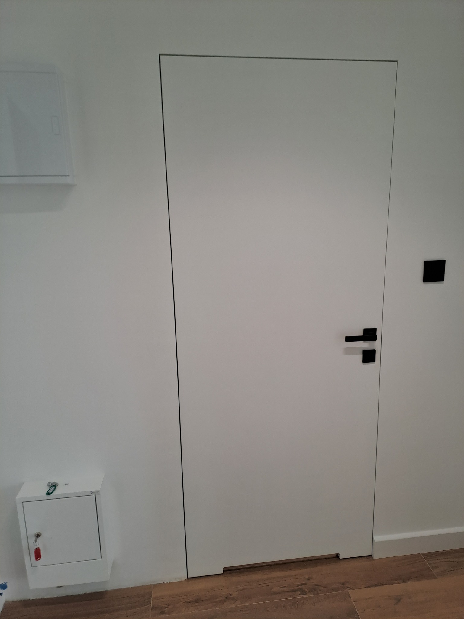 Białe, minimalistyczne drzwi z czarną klamką i ukrytą ościeżnicą, zamontowane w świeżo odmalowanej, białej ścianie, z widoczną skrzynką z kluczem poniżej i brązową podłogą z płytek imitujących drewno.