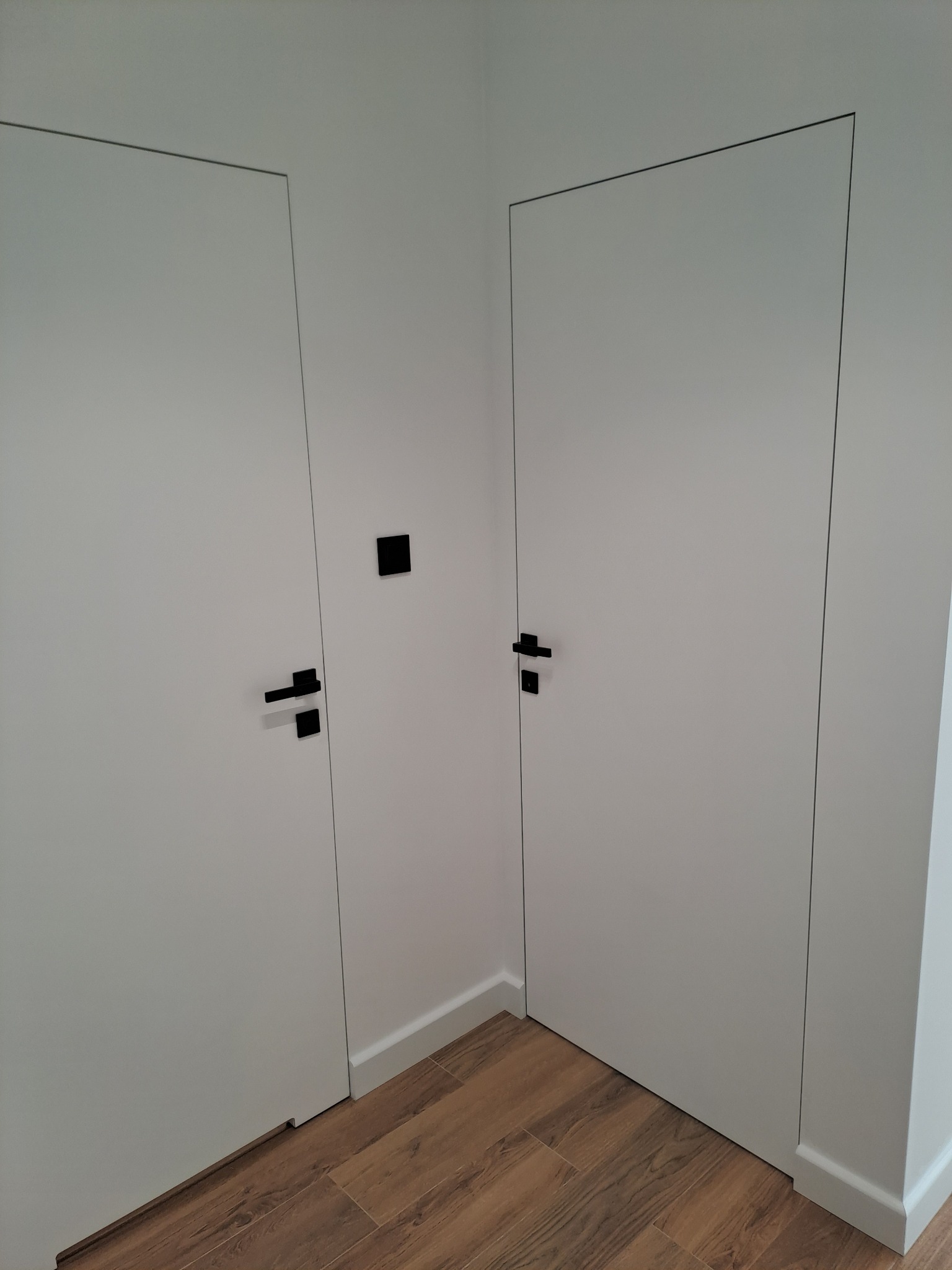 Dwa białe, minimalistyczne drzwi wewnętrzne z czarnymi klamkami, ukryte w ścianie, widoczne na tle drewnopodobnej podłogi i białych listew przypodłogowych.