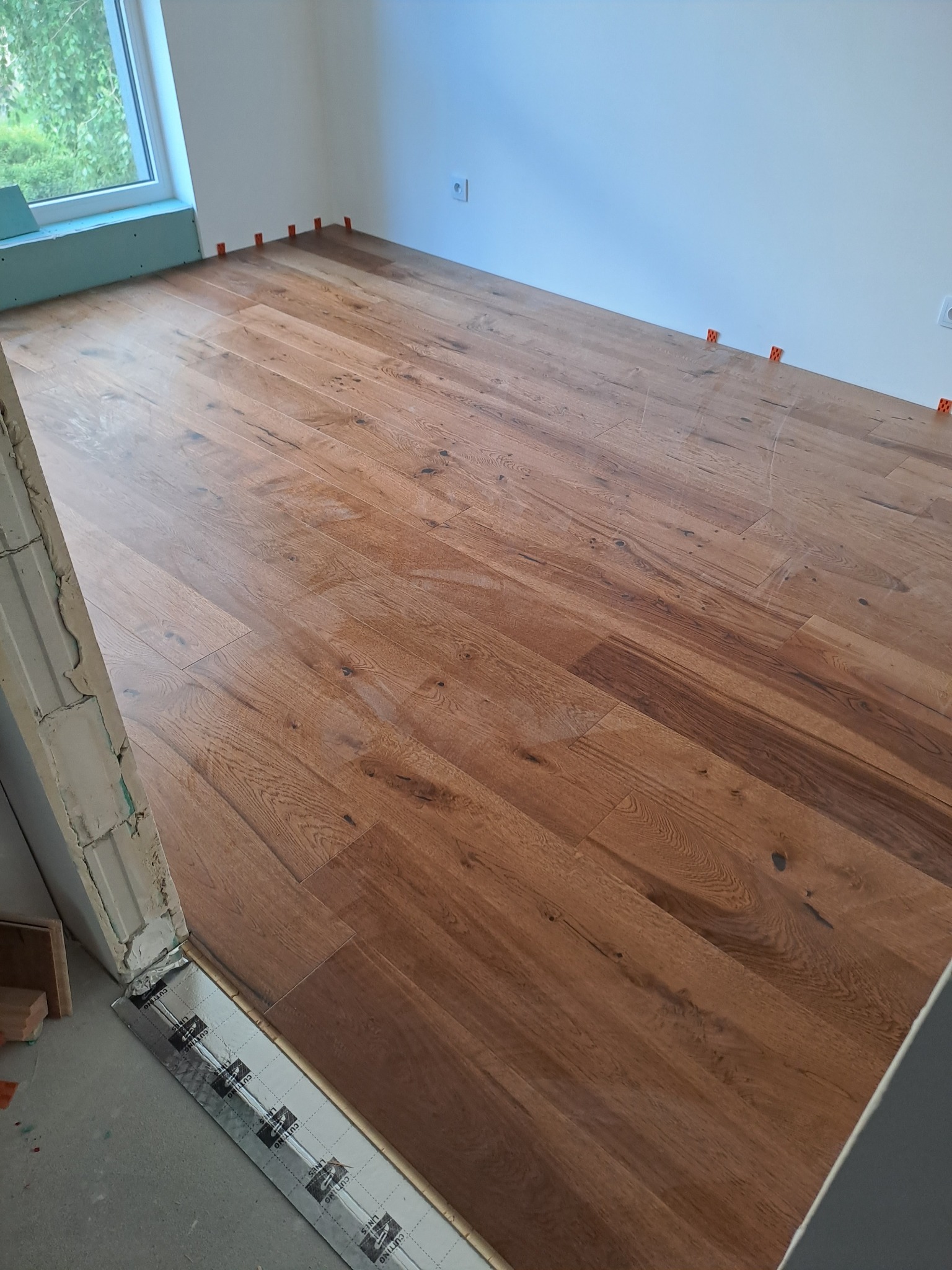 Układanie drewnianej podłogi w jasnym pomieszczeniu, widoczne pomarańczowe kliny dystansowe przy ścianach, fragment surowej ściany przy wejściu z izolacją podłogową.