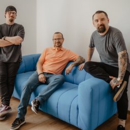 Blendify AI - Trzech mężczyzn w swobodnych ubraniach pozuje na tle białej ściany i niebieskiej sofy. Jeden stoi z założonymi rękami, drugi siedzi, a trzeci opiera się o sofę.