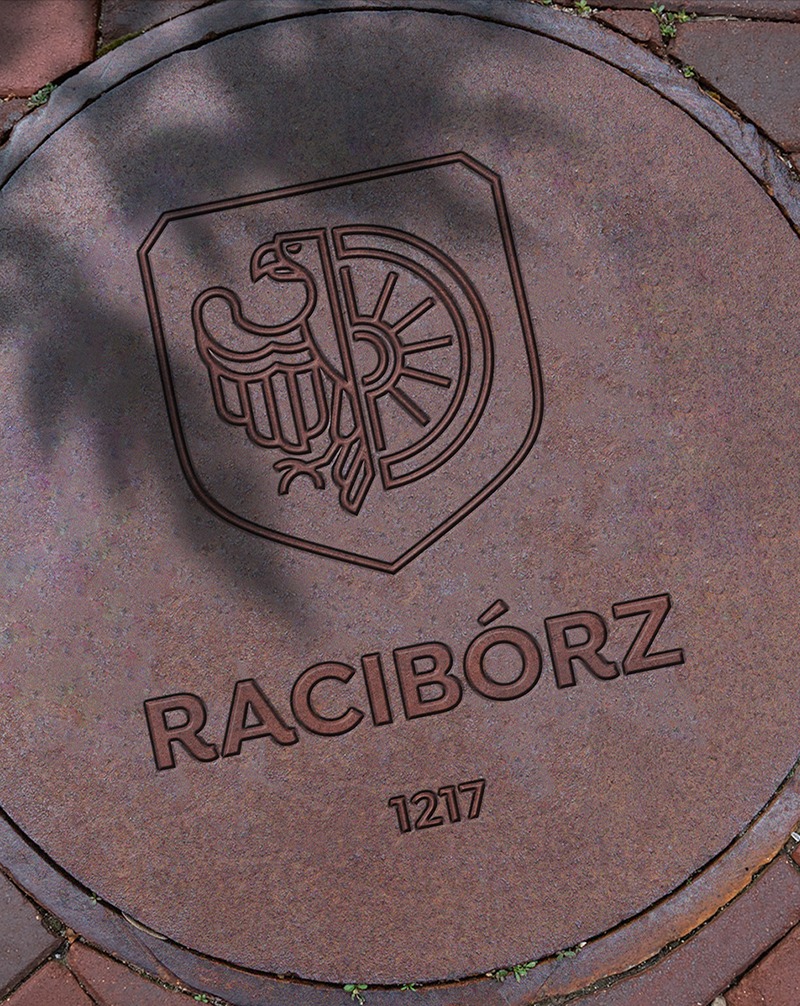 Metalowa pokrywa studzienki z wygrawerowanym herbem Raciborza (orzeł i koło) oraz rokiem 1217, otoczona kostką brukową i zielenią. Ciemne cieniowanie.