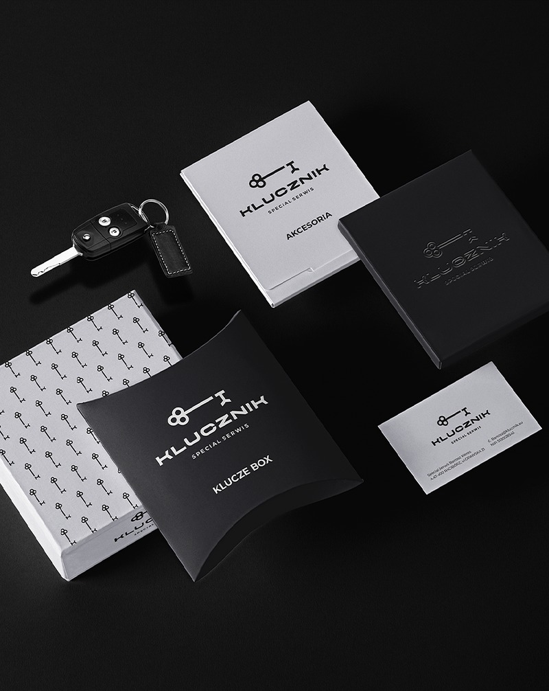Elegancki zestaw identyfikacji wizualnej firmy Klucznik: pudełka, wizytówka i kluczyk samochodowy na czarnym tle. Minimalistyczny design w bieli i czerni.
