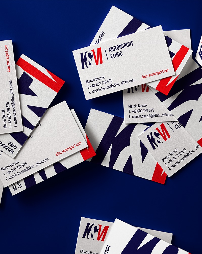 Stos wizytówek K&M Motorsport Clinic na granatowym tle. Minimalistyczny design z czerwonymi i granatowymi akcentami. Widoczne dane kontaktowe i logo firmy.