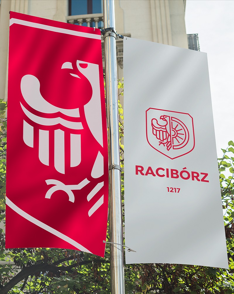 Dwa pionowe banery z herbem i nazwą Racibórz (1217) na tle zieleni i budynku. Czerwony baner z białym orłem, biały baner z czerwonym herbem i napisem.