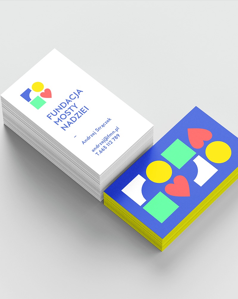 Dwa stosy wizytówek: białe z logo Fundacji Mosty Nadziei i niebieskie z abstrakcyjnym wzorem geometrycznym. Żółte krawędzie wizytówek dodają koloru. Minimalistyczny design.
