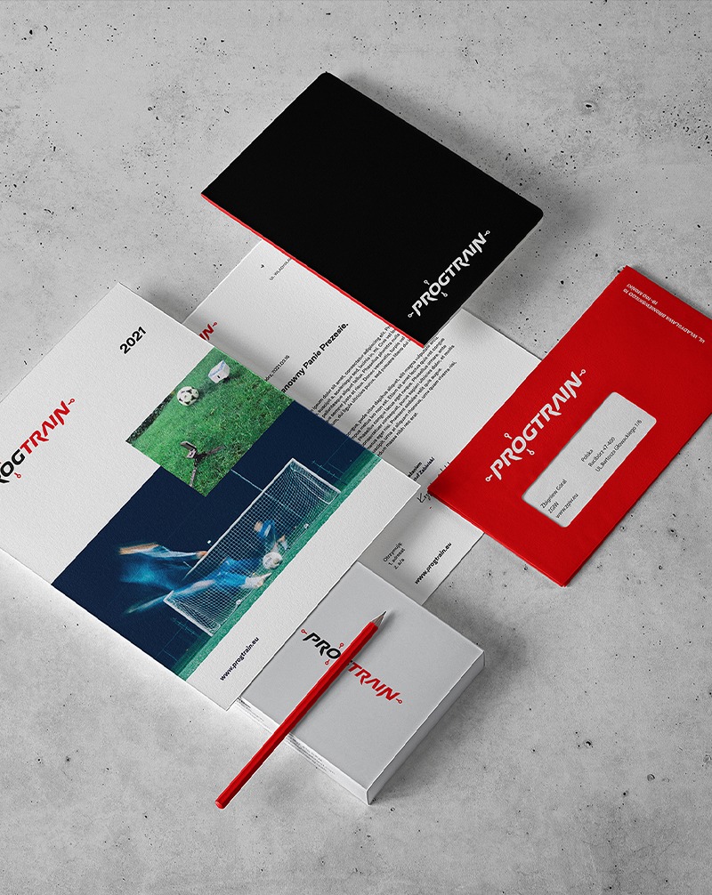 Zestaw materiałów firmowych z logo: wizytówki, koperta, teczka, papier firmowy. Czerwono-czarna kolorystyka, nowoczesny design. Widok z góry na szarym tle.