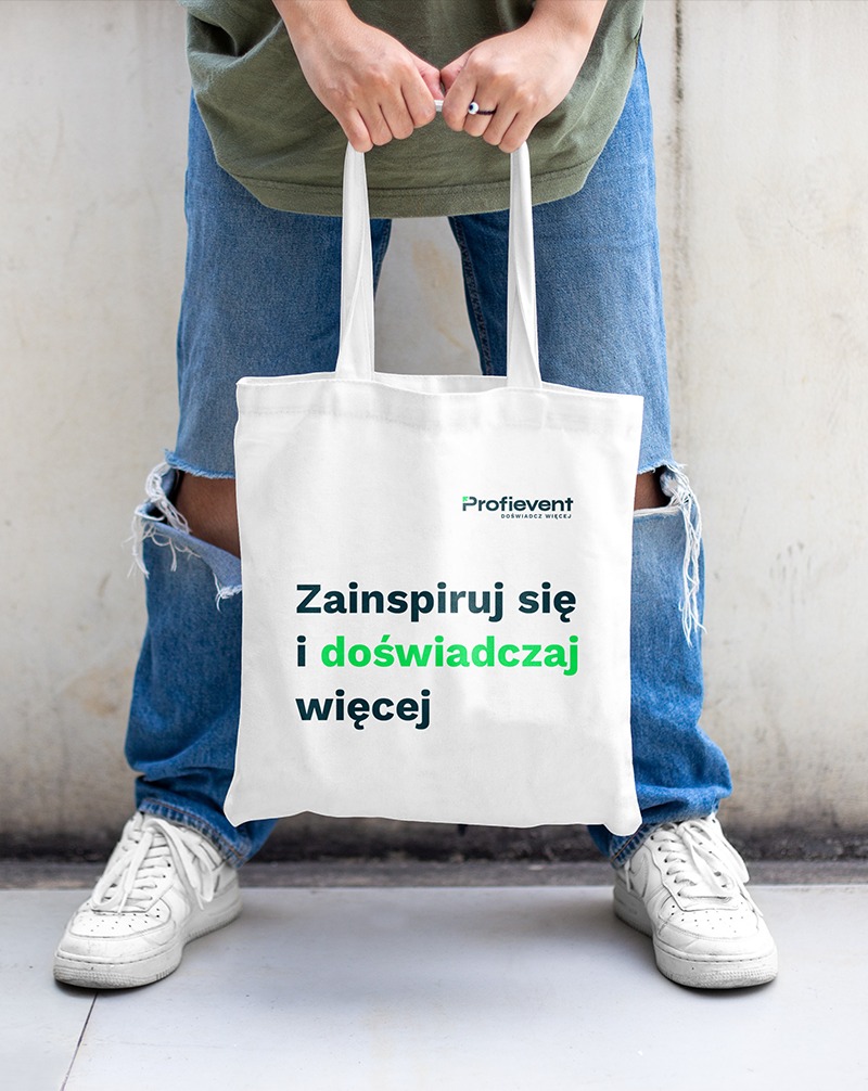 Osoba trzyma białą torbę z logo Profievent i hasłem 'Zainspiruj się i doświadczaj więcej'. Widoczne nogi w jeansach i białych butach. Minimalistyczny design torby.