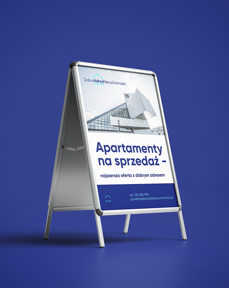 Stojak reklamowy A-frame z ofertą sprzedaży apartamentów, logo 'Dobry Adres Nieruchomości' i nowoczesną architekturą w tle, na jednolitym, niebieskim tle.