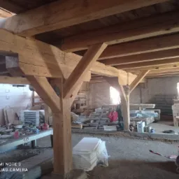 Wnętrze stodoły z drewnianą konstrukcją nośną, widoczne belki stropowe, słupy i podciągi, w tle materiały budowlane i narzędzia pracy, naturalne oświetlenie wpadające przez okno.