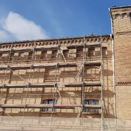 Wielopiętrowy budynek z cegły w trakcie renowacji, otoczony rusztowaniami, widoczne detale architektoniczne i fragment niebieskiego nieba.