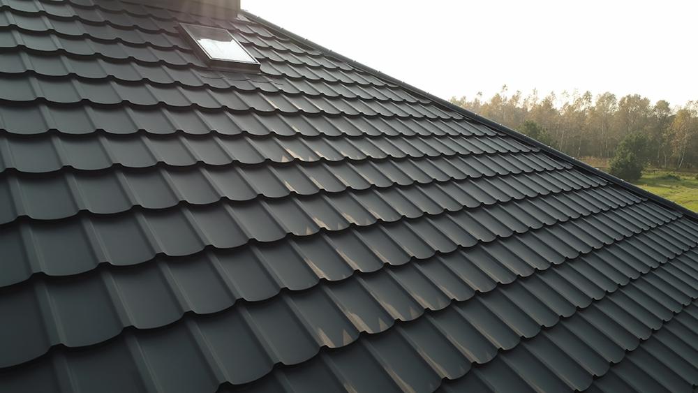 Nowy, grafitowy dach z blachodachówki modułowej z oknem dachowym, widok z dołu pod kątem, las w tle.
