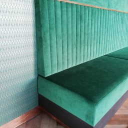 Łukasz Starosta HEST MEBLE TAPICEROWANE - Szmaragdowa sofa tapicerowana z pionowymi przeszyciami, drewnianym akcentem i wzorzystą tapetą w tle. Ujęcie z perspektywy narożnika, podkreślające detale wykończenia.