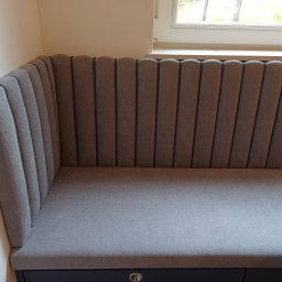 Łukasz Starosta HEST MEBLE TAPICEROWANE - Tapicerowany narożnik wnękowy w odcieniach szarości, z pionowymi przeszyciami na oparciu, umieszczony przy oknie. Widoczny fragment siedziska i oparcia.